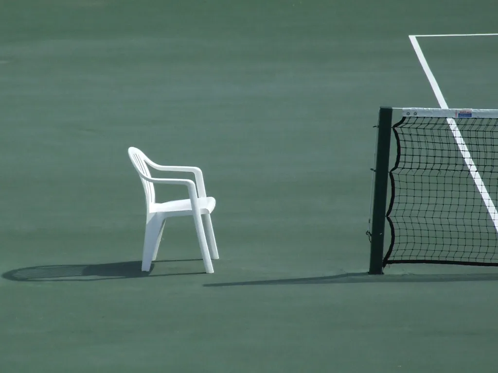 Silla de plástico vacía junto al poste de una red de tenis en una pista sin jugadores